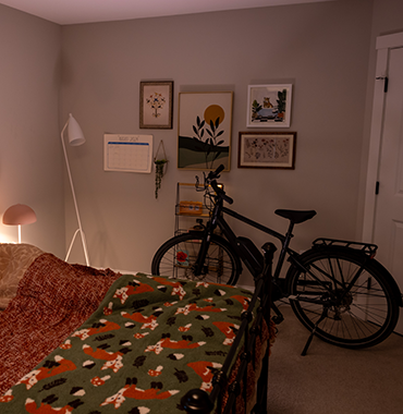 A cozy bedroom with a bike leaning against the wall. Framed art and a calendar hang above the bike. A bed with a green and orange patterned blanket is on the left and a standing lamp with a pink shade is beside it. A closed white door is visible to the right.