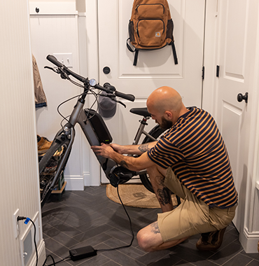 A person with a bald head and a beard, wearing a striped shirt and shorts, is crouching to charge an electric bike inside a small room. A brown backpack hangs on the wall, and a wooden floor with a mat is visible.