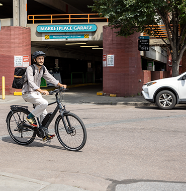 A person wearing a helmet and carrying a backpack rides an electric bicycle past an entrance to the Marketplace Garage in an urban setting. A white car and some trees are visible in the background.