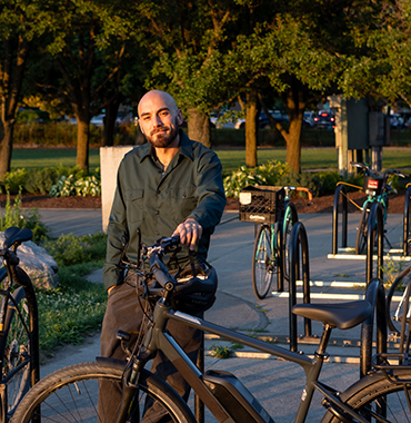 A person with a beard is standing outdoors next to a black bicycle at a bike rack. They are wearing a long-sleeved dark shirt and pants. In the background, there are trees, parked bicycles, and a road with a few cars. The sun is low, casting a warm light.