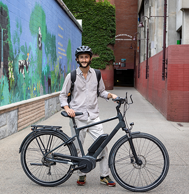 A person in a helmet stands next to a black bicycle in a narrow alleyway. They are smiling, wearing a long-sleeved shirt and light-colored pants, and have a backpack. The alley has a vibrant mural on one side and red brick walls on the other.