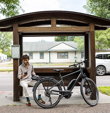 A man sits at a wooden bus stop shelter, focused on his smartphone. His bicycle is parked in front of the shelter. A passing white vehicle and houses are visible in the background. The scene is outdoors on a clear day.