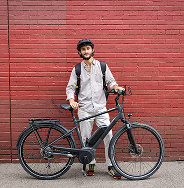 A man with a beard wearing a grey shirt, matching pants, and a black helmet stands outside against a red brick wall, smiling and holding a black bicycle. He has a black backpack on his shoulders.