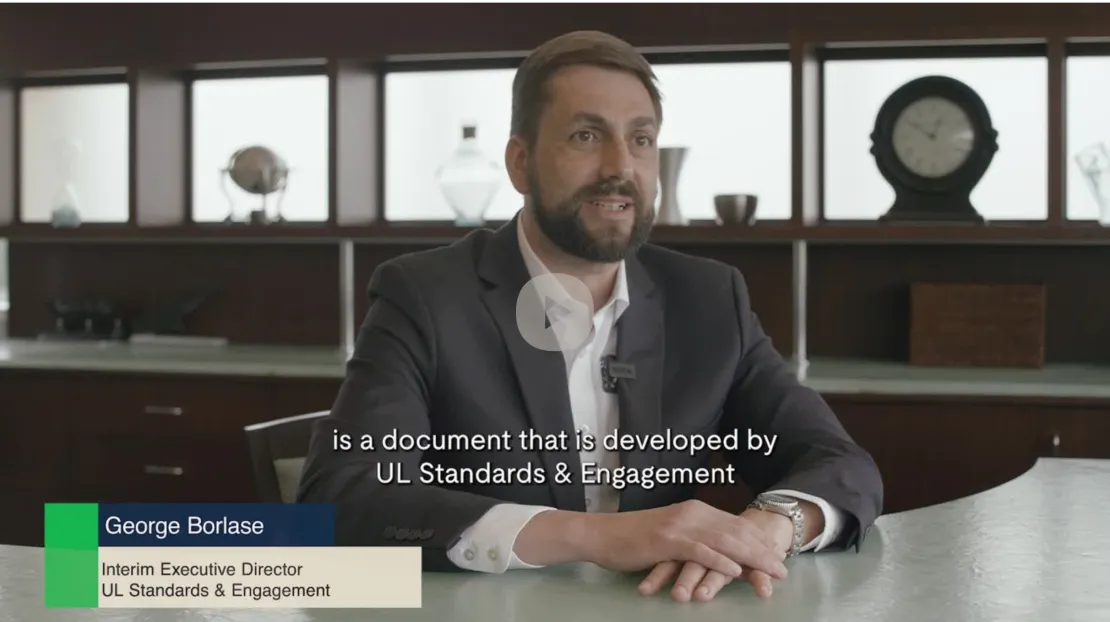 A man in a suit, identified as George Borlase, Interim Executive Director of UL Standards & Engagement, is speaking in an office setting. Subtitles on the image read: "is a document that is developed by UL Standards & Engagement.