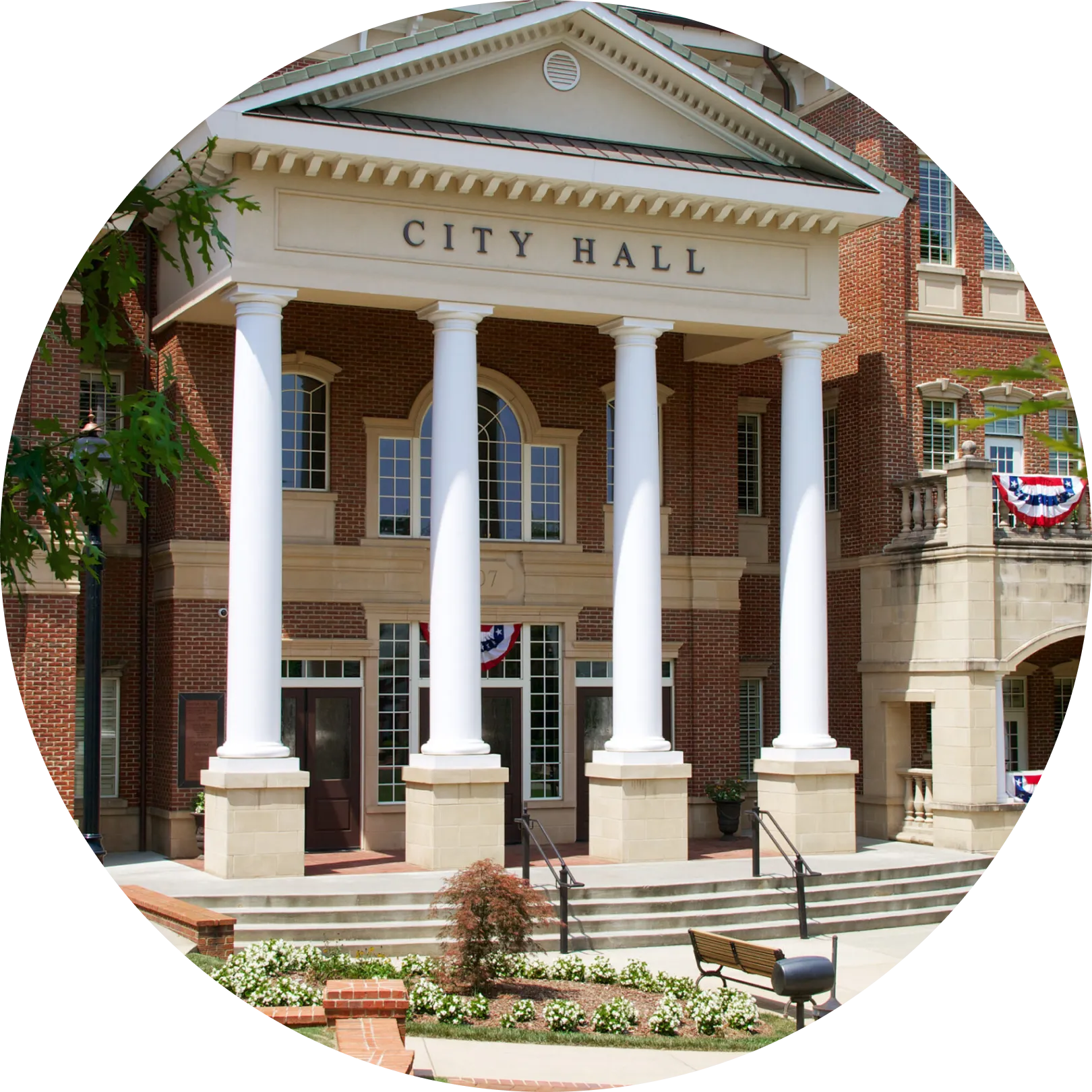 A brick city hall building with tall white columns and an arched entrance. "City Hall" is inscribed above the entrance. Patriotic red, white, and blue bunting decorates the building. Steps lead up to the entrance, and flowers and a bench are in the foreground.