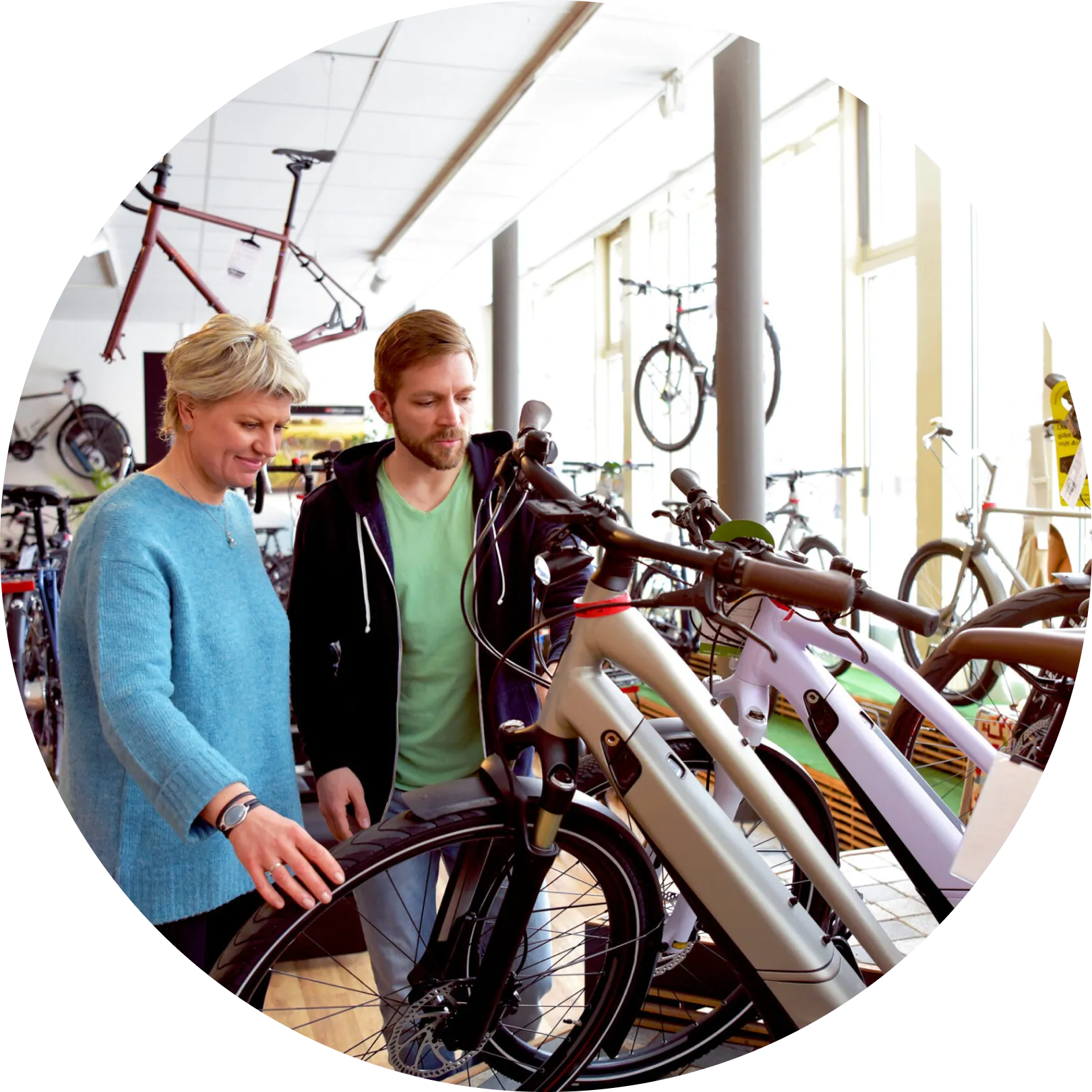 Two people, a woman in a blue sweater and a man in a green T-shirt with a black hoodie, are looking at bicycles in a bike shop. Bicycles are displayed around the store, some hanging on the walls and others on the floor. The shop has large windows with natural light.