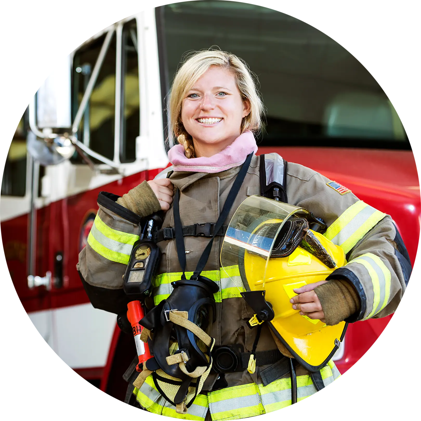 A firefighter in full gear smiles while standing in front of a fire truck. She holds a yellow helmet in one hand and has various equipment strapped to her suit. The fire truck behind her is red and white.